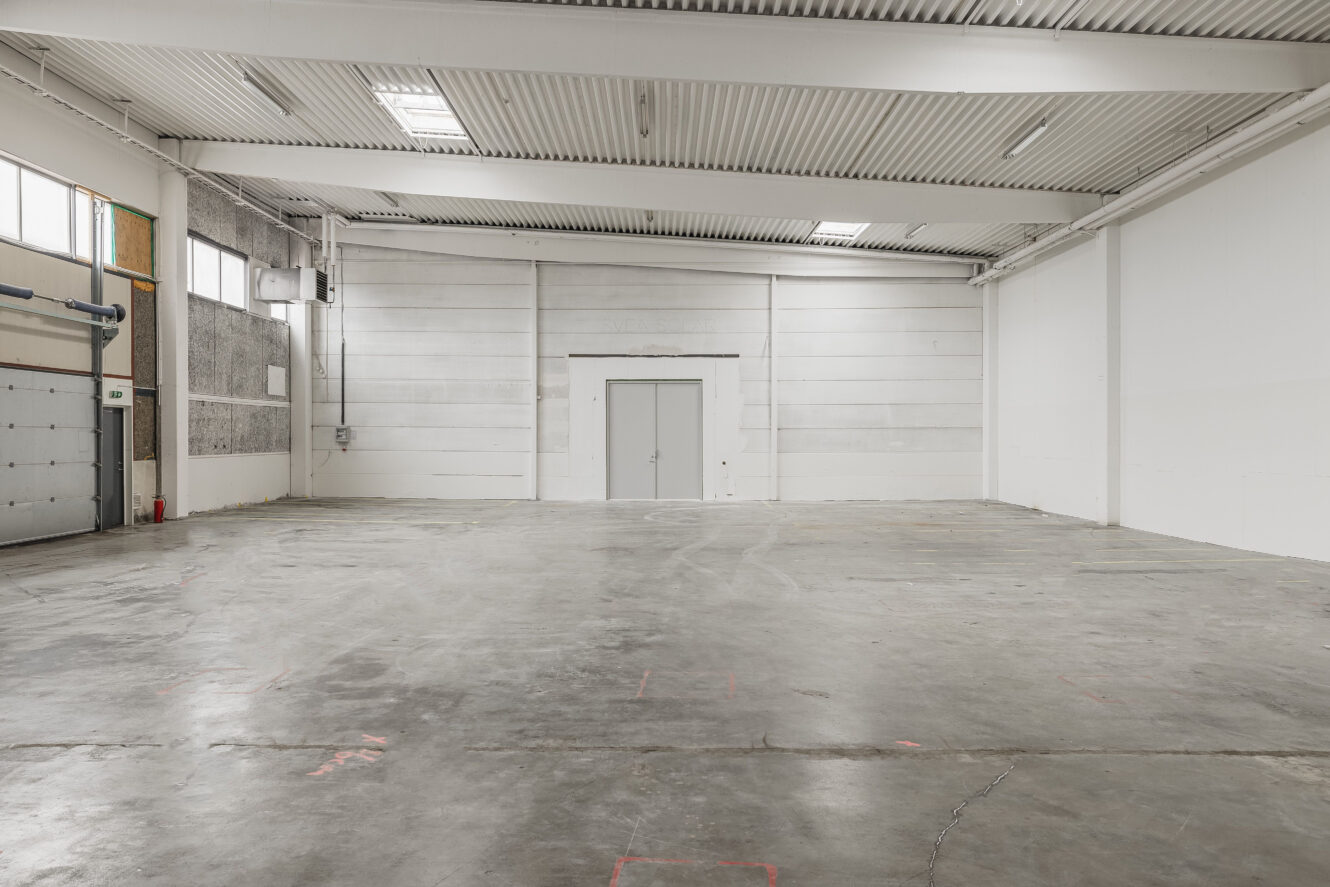 Stor, tom industrilagerlokal med betonggolv, vita väggar, högt i tak och stängda dubbeldörrar i bortre änden.