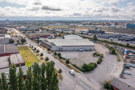 Flygfoto över ett industriområde med lagerbyggnader, parkerade lastbilar, vägar och lite grönska under en delvis molnig himmel.