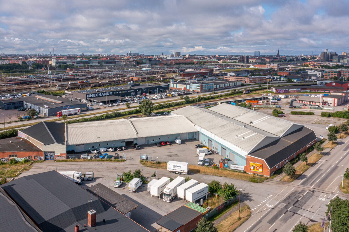 Flygfoto över ett industriområde med lagerlokaler, lastbilar parkerade utanför och järnvägsspår i bakgrunden under en delvis molnig himmel.