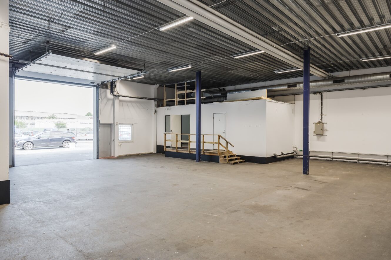 Rymlig industriell lagerlokal med betonggolv, öppen garageport, taklampor och vitt kontorsutrymme på en upphöjd plattform med trappor. Bilar synliga utanför.