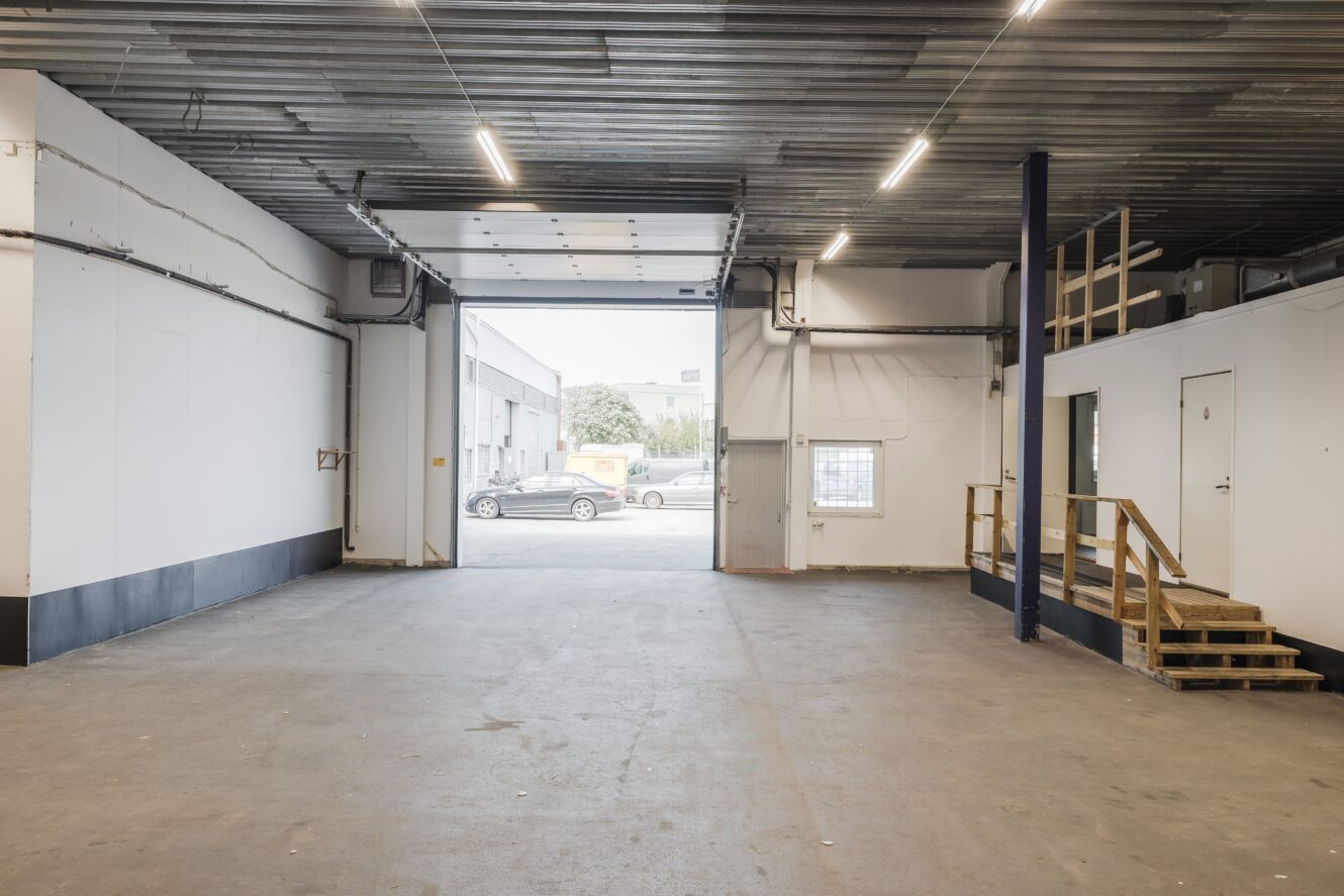 Tom lagerlokal med betonggolv, högt i tak, öppen garageport, trätrappa som leder till ett litet entresolplan och utsikt över parkerade bilar och byggnader utanför.