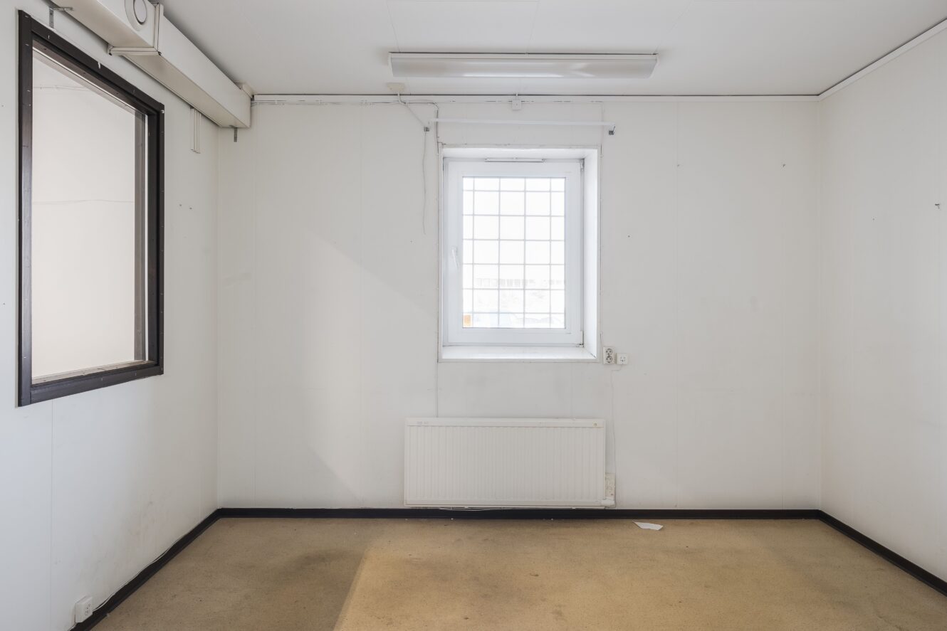 Tomt vitt rum med ett enda fönster, ett element under fönstret, beige heltäckningsmatta och ett svartmålat innerfönster på vänster vägg.