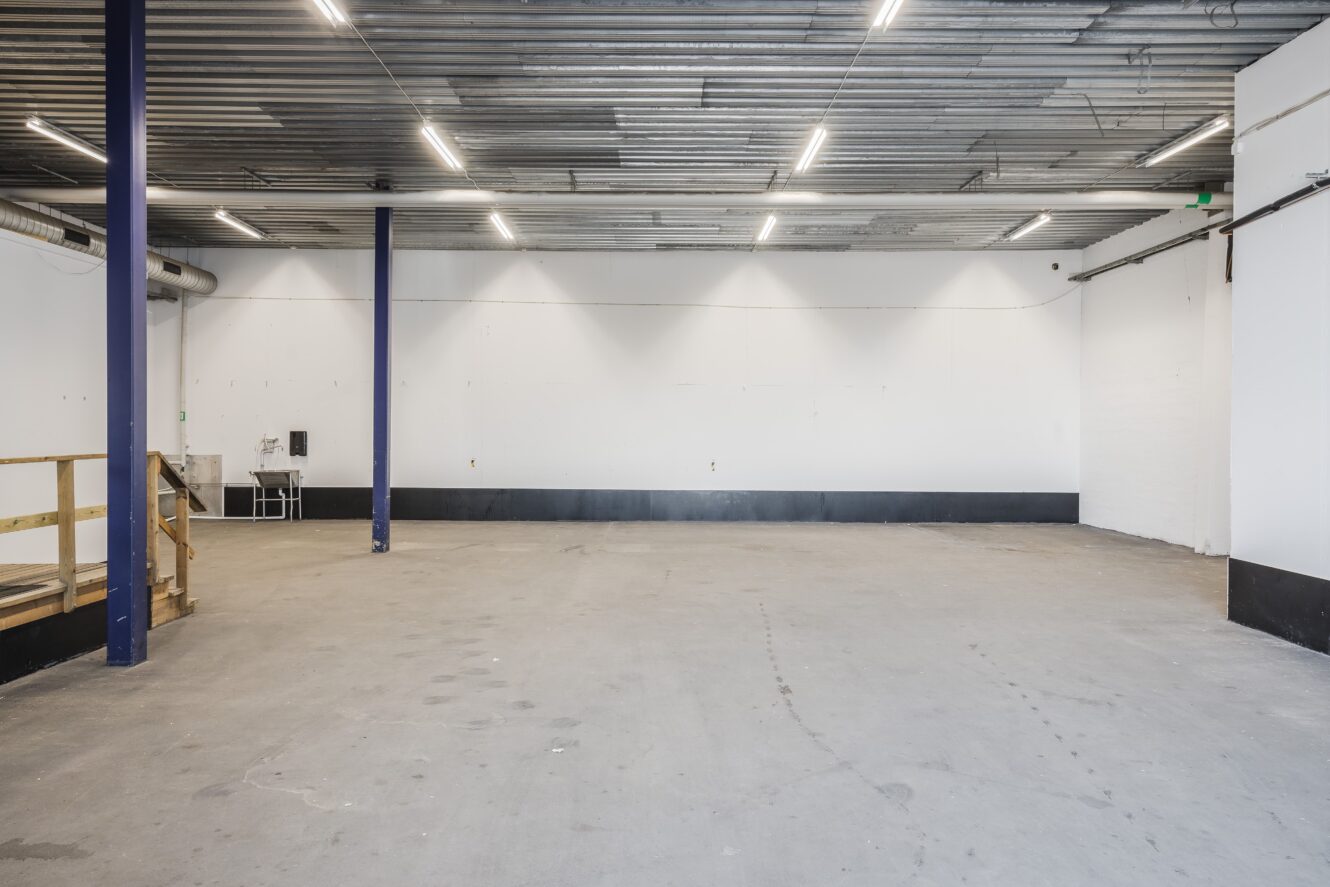 Tomt industrilager med betonggolv, vita väggar, synligt tak, lysrörsbelysning och en liten diskho i hörnet.