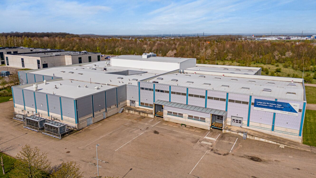 En stor industribyggnad med ett platt vitt tak och blå accenter, omgiven av en asfalterad tomt och grönska, en klar dag.