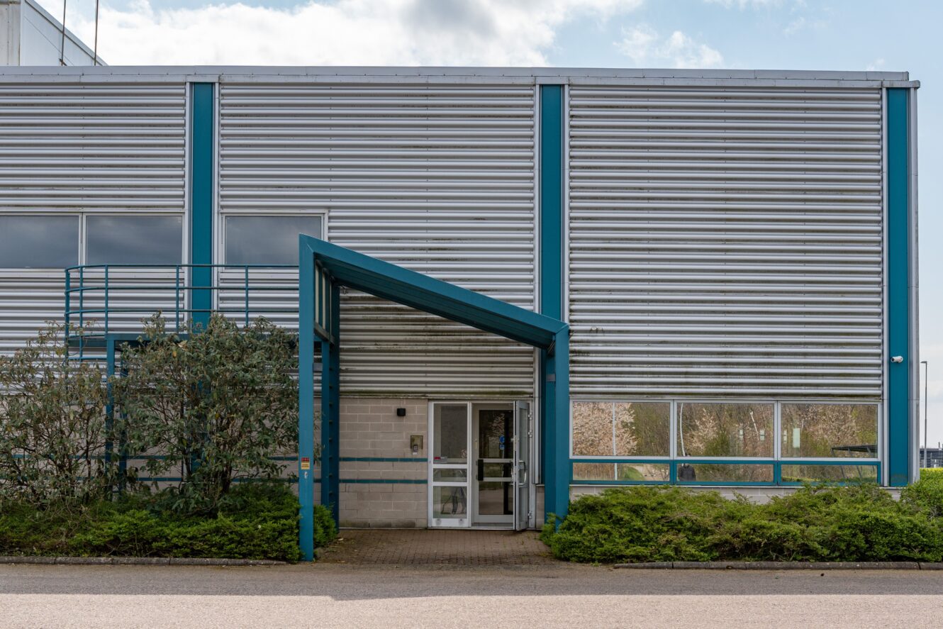 En kommersiell eller industriell byggnad med en metallfasad, blå strukturella accenter, stora fönster och en täckt glasentré omgiven av buskar.