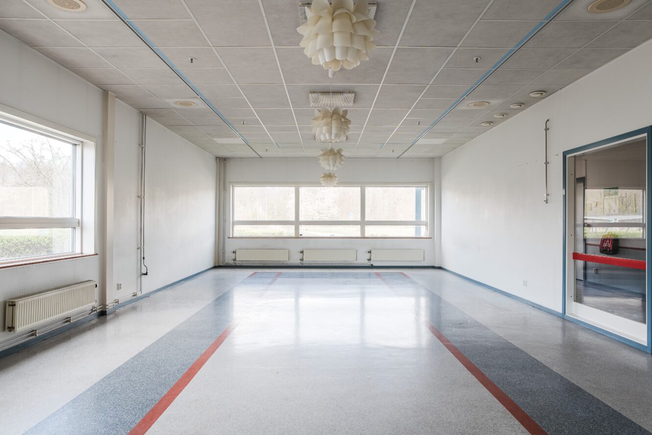 Ett tomt rum med stora fönster, tre taklampor, vita väggar, grått golv och röda och blå golvlinjer.