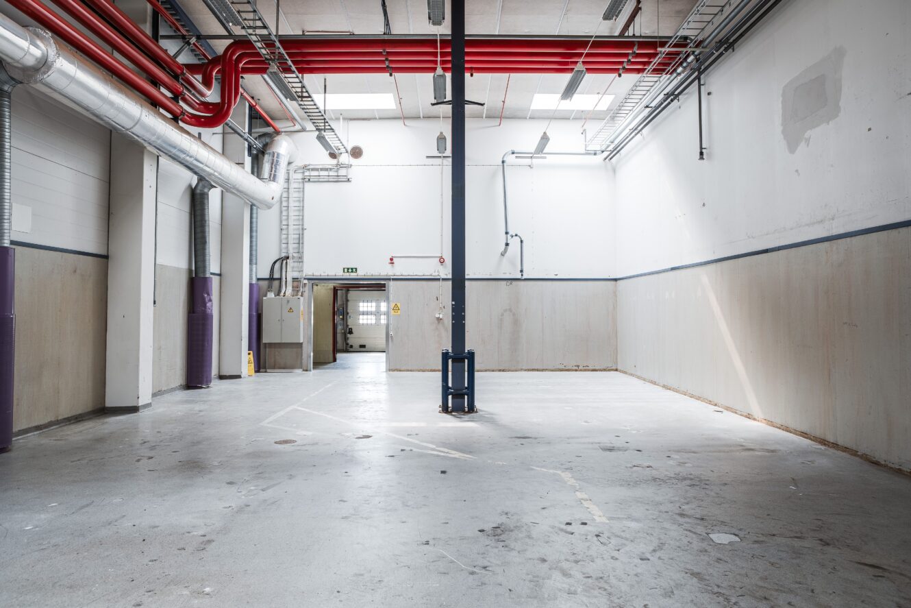Tomt industrilager med betonggolv, högt i tak, synliga rör och en central stödpelare. Ljusinsläpp genom fönster och en dörr på baksidan.