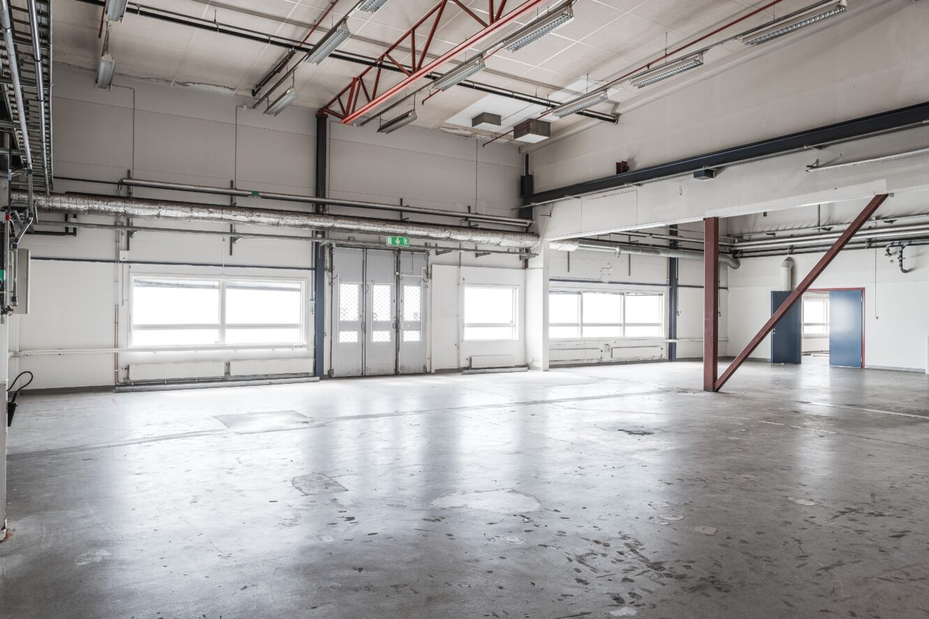 Tomt industrilager med betonggolv, stora fönster, synliga rör, konstruktionsbalkar och takbelysning.
