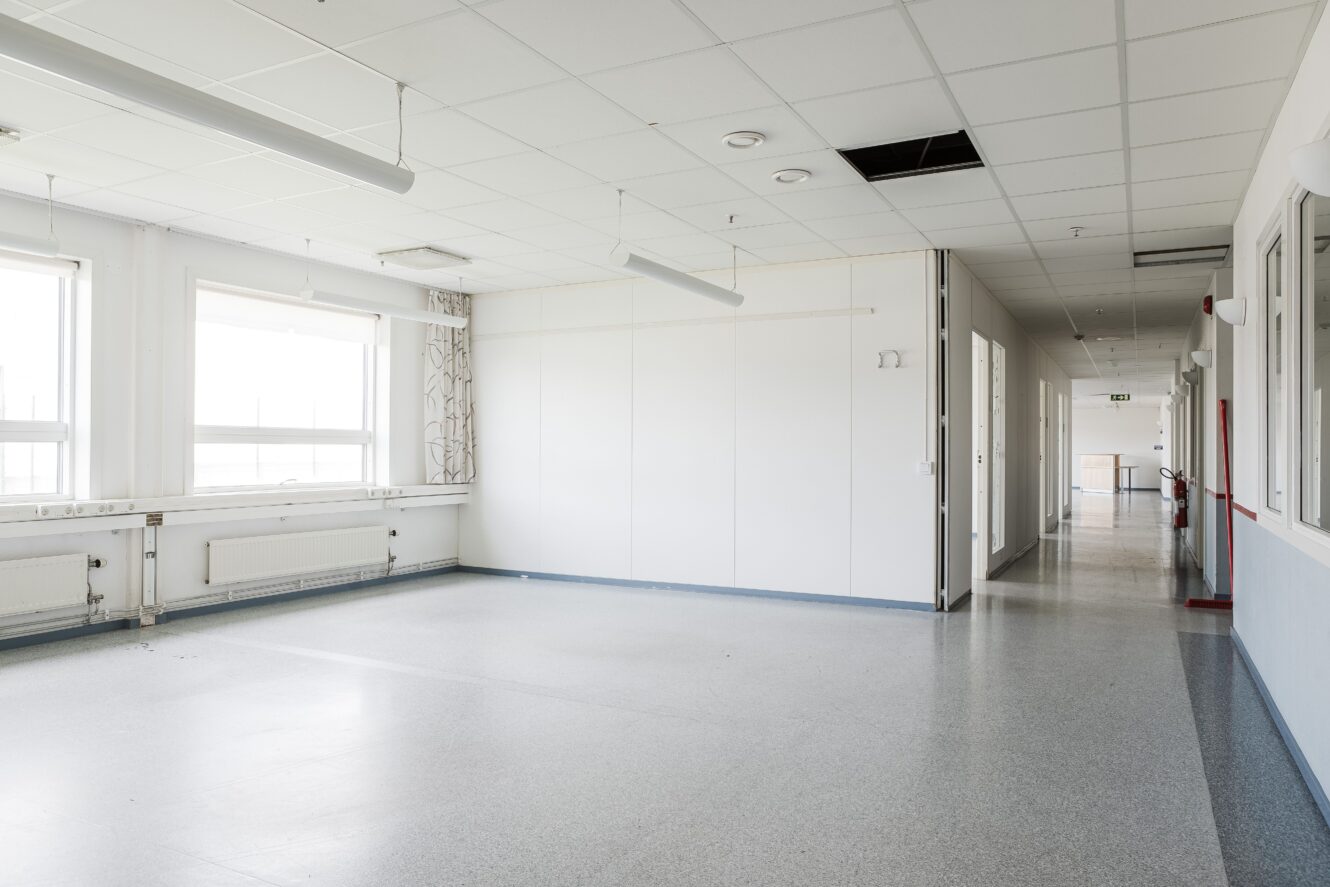 Tomt sjukhusrum med stora fönster, vita väggar och kaklat tak, i anslutning till en korridor; en takplatta saknas.