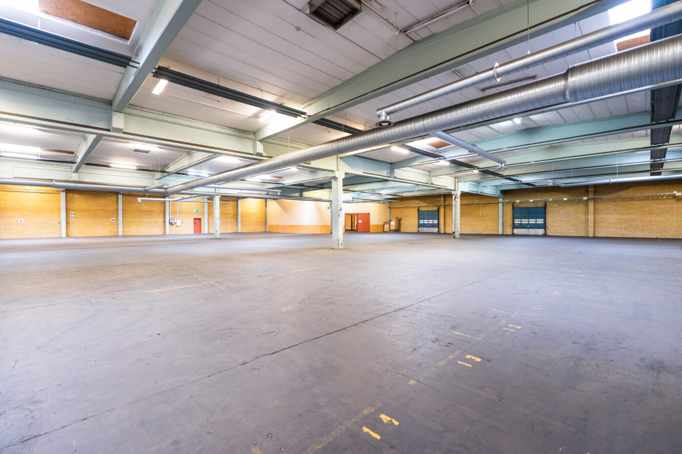 Stor tom lagerlokal med högt i tak, synliga bjälkar, industriell belysning och gula tegelväggar. Golvet är av betong och markerat med målade linjer och siffror.