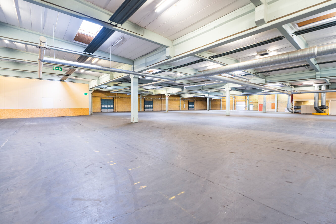 Rymlig, tom industrilagerinredning med betonggolv, högt i tak, synliga bjälkar och stora garageportar längs väggarna.