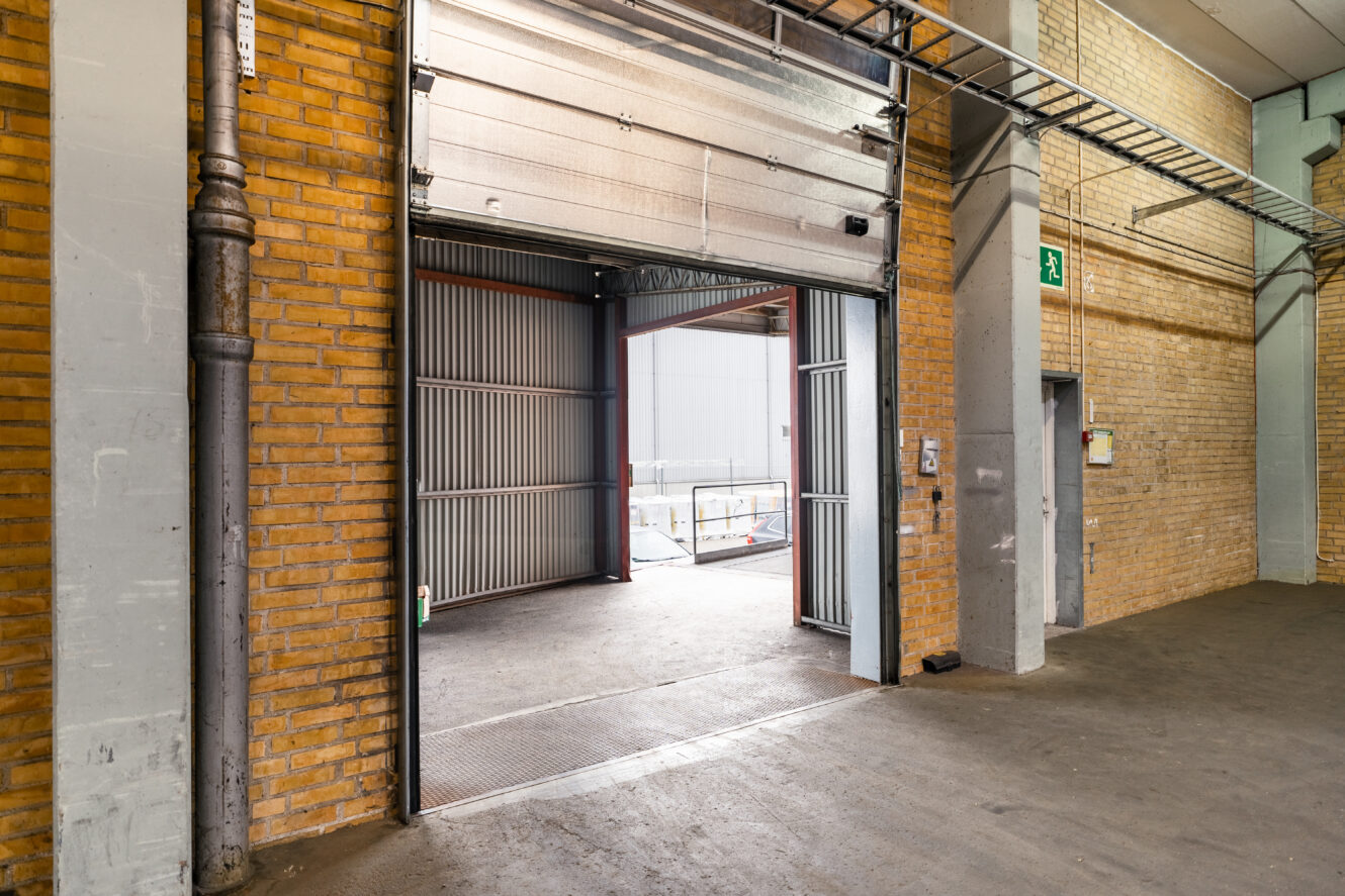 Öppen lastkaj för industriellt lager med tegelväggar, betonggolv och en delvis upphöjd garageport i metall som leder till en utvändig serviceplats.