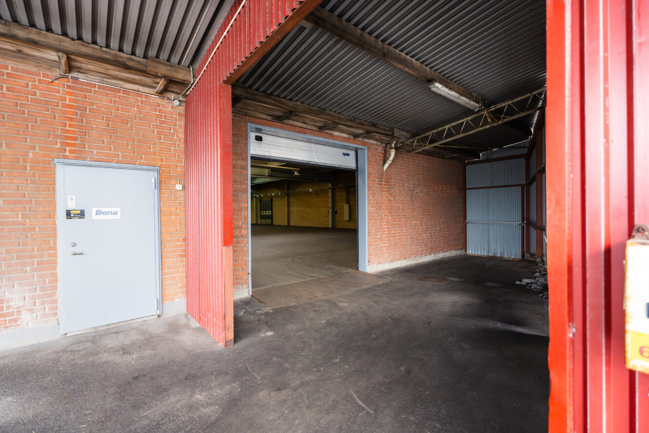 Ett stort industriellt lager med röda tegelväggar, en grå dörr märkt Bona och en öppen rullport som leder till en rymlig, tom interiör.