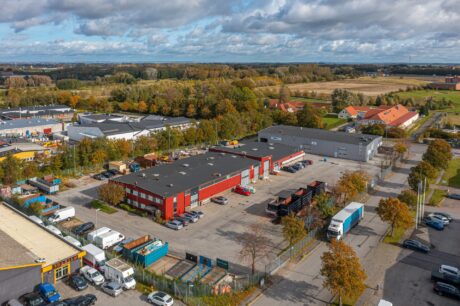 Flygfoto över ett industriområde med lagerbyggnader, lastbilar och parkerade fordon omgivna av träd och närliggande öppna fält under en delvis molnig himmel.