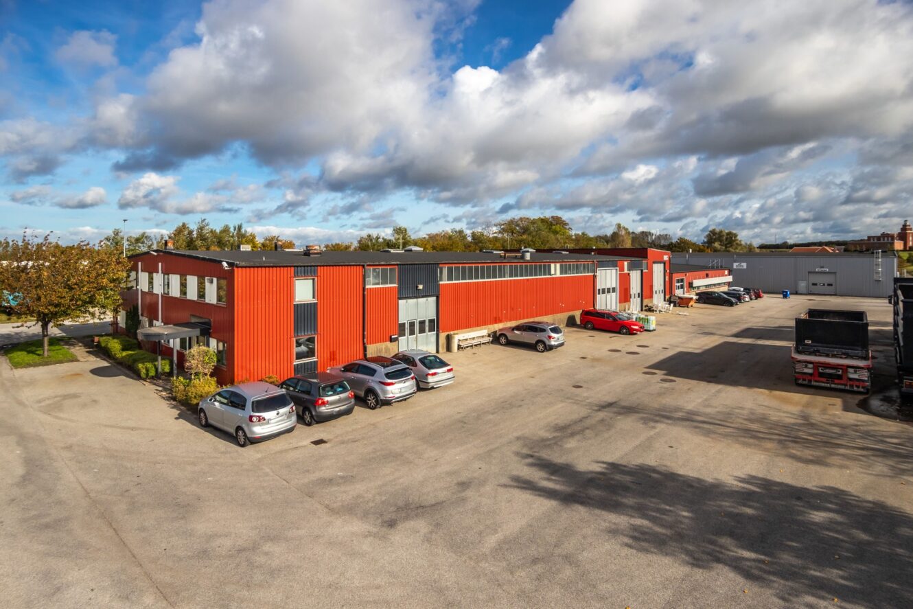 En rad röda industribyggnader med parkerade bilar och lastbilar på en stor öppen tomt under en delvis molnig himmel.