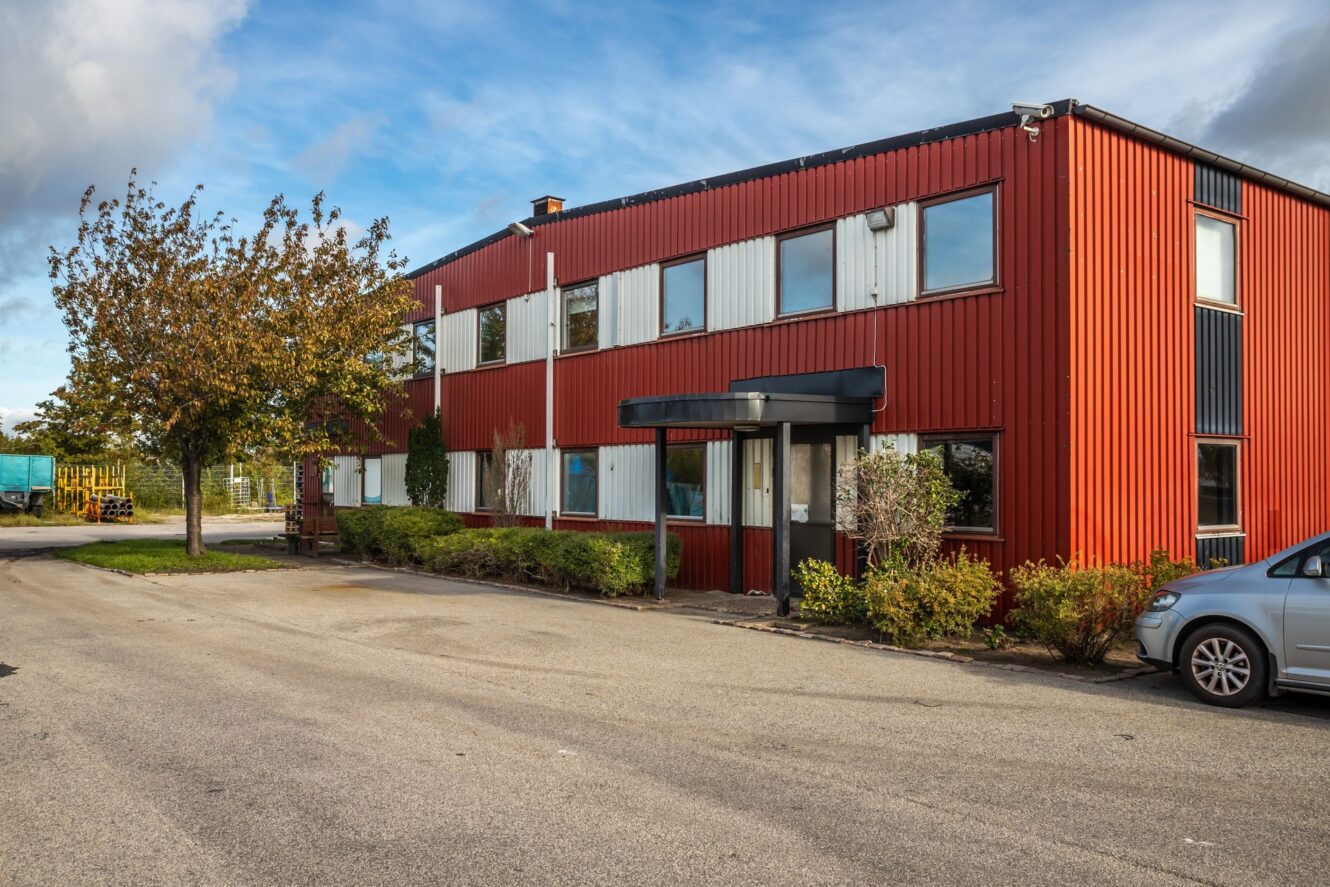 En röd industribyggnad i två våningar med vita fönsterkarmar, en svart markis över entrén och ett träd och buskar framför, på en asfalterad tomt under en delvis molnig himmel.