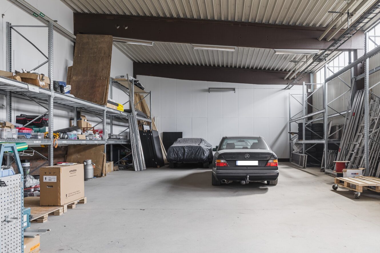 Ett rymligt garage med två parkerade bilar, varav en täckt, omgiven av metallhyllor med olika verktyg, lådor och material.