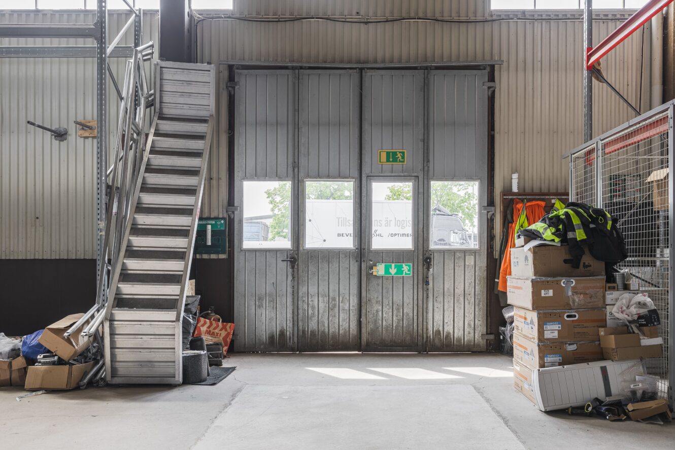 Industriell lagerinredning med metalltrappor till vänster, lådor och säkerhetsutrustning till höger och en stor dubbeldörr i metall med utgångsskyltar i mitten.