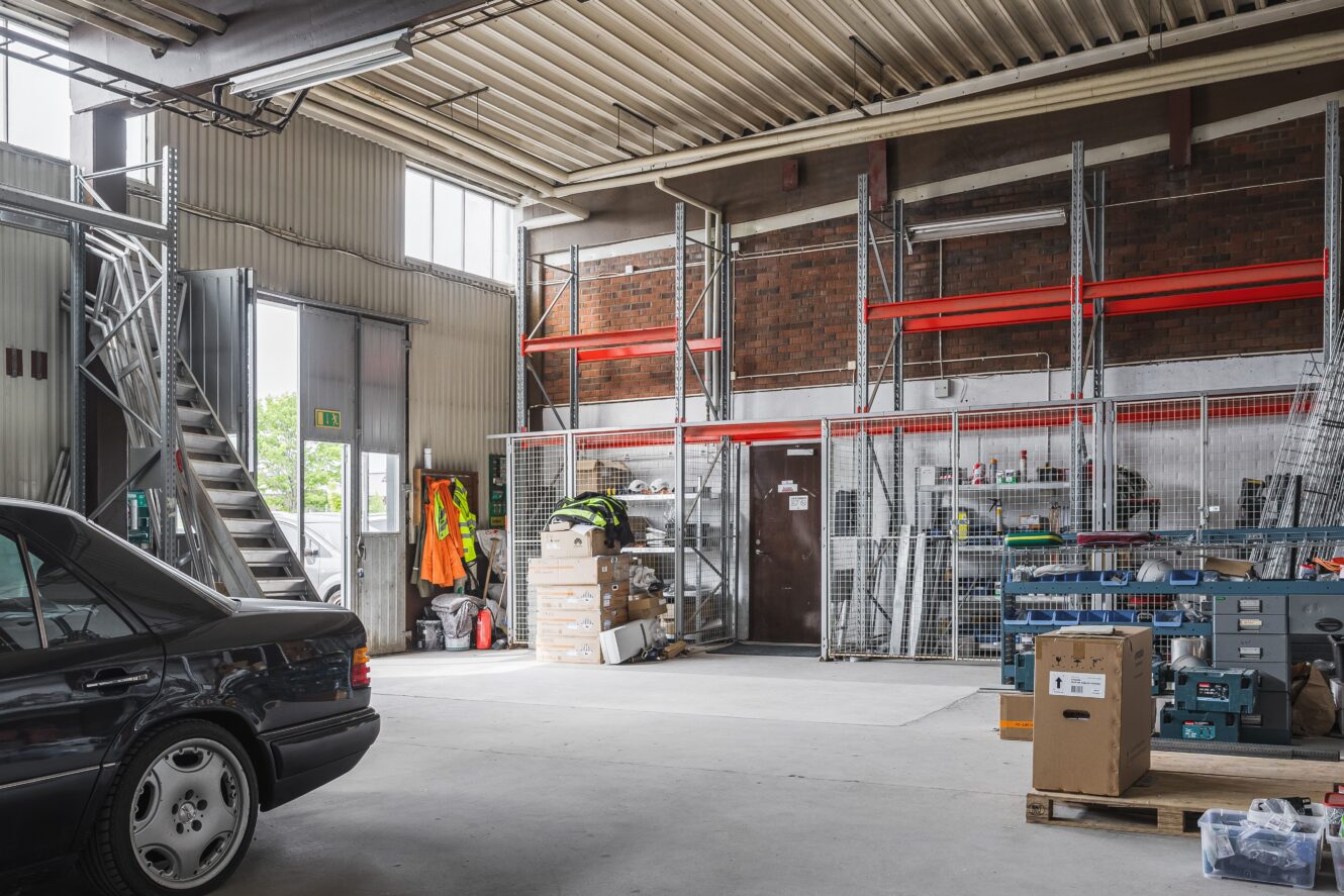 Inredning i ett industriellt garage eller lager med hyllor, förvaringslådor, verktyg, en svart bil och höga fönster som släpper in naturligt ljus.