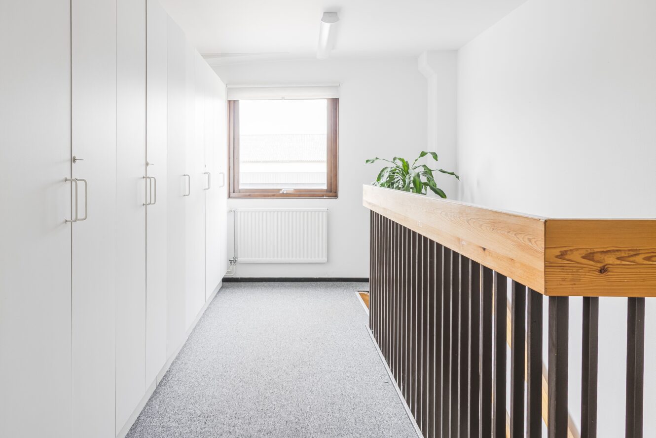 En ljus hall med vita skåp till vänster, ett fönster med en radiator under, en krukväxt och ett träräcke till höger.