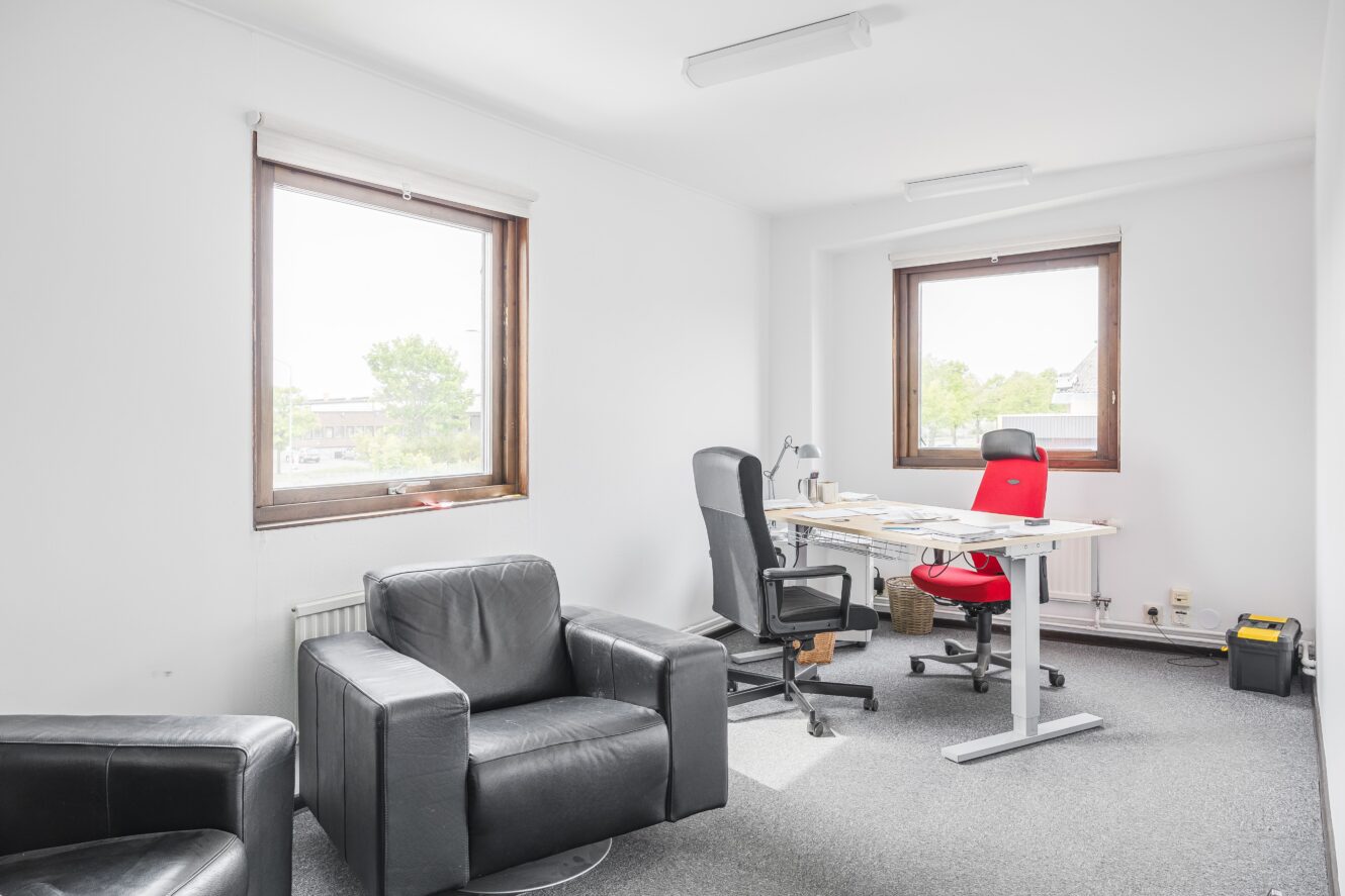 Ett litet kontor med två svarta fåtöljer, ett skrivbord med två kontorsstolar - en svart och en röd - och två fönster som släpper in naturligt ljus.