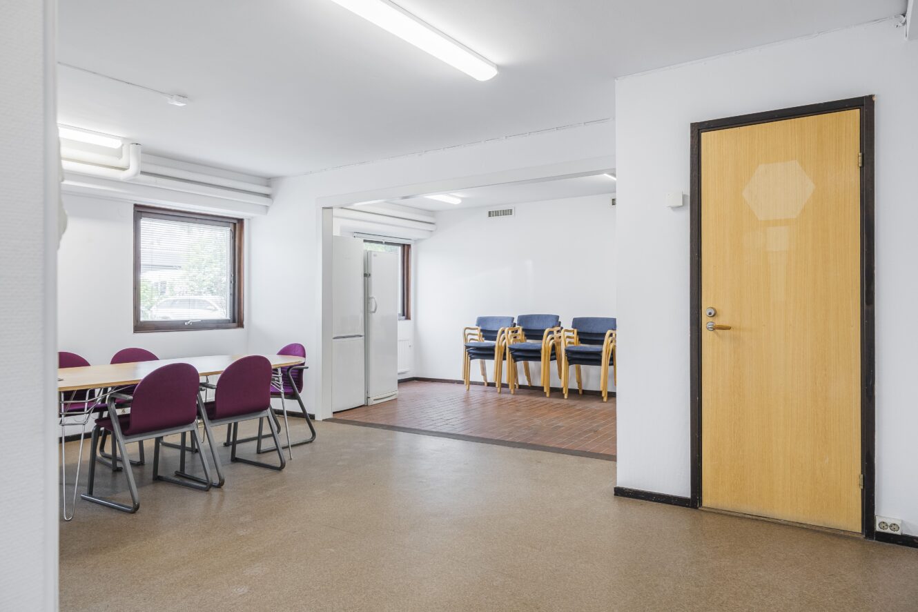 Ett rent, enkelt rum med bord och stolar, en dörr, fönster och staplade stolar i hörnet nära ett kylskåp.