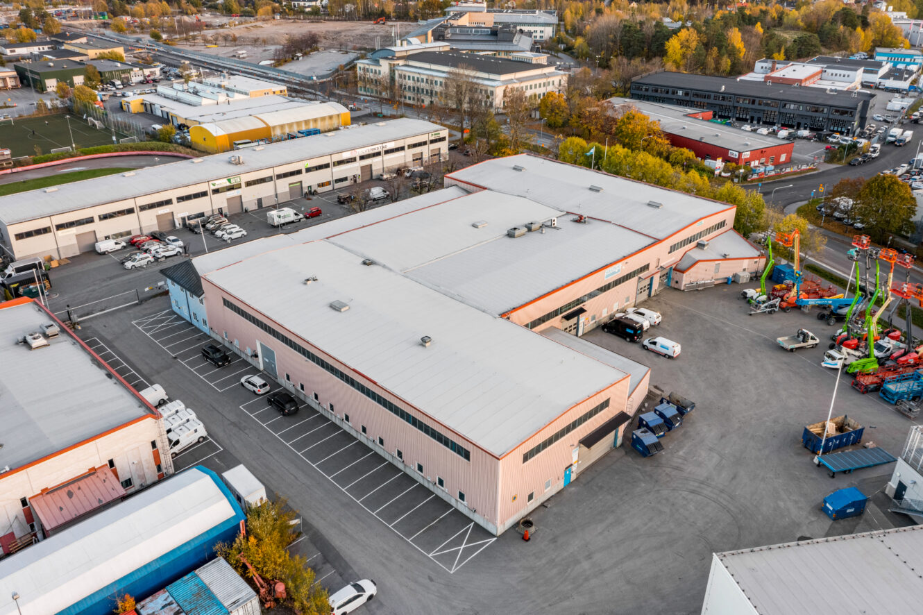 Flygfoto över en stor industrihall med vitt tak, omgiven av parkerade fordon och intilliggande byggnader i ett affärsområde.