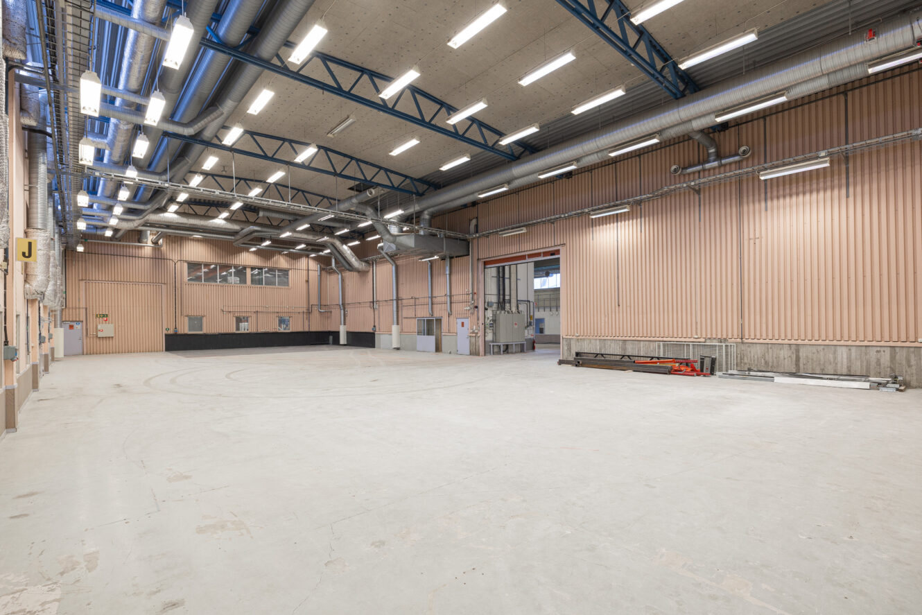 Stort, tomt industrilager med högt i tak, synliga ventilationskanaler, beige väggar och stark takbelysning. En del utrustning är staplad längs den högra väggen.