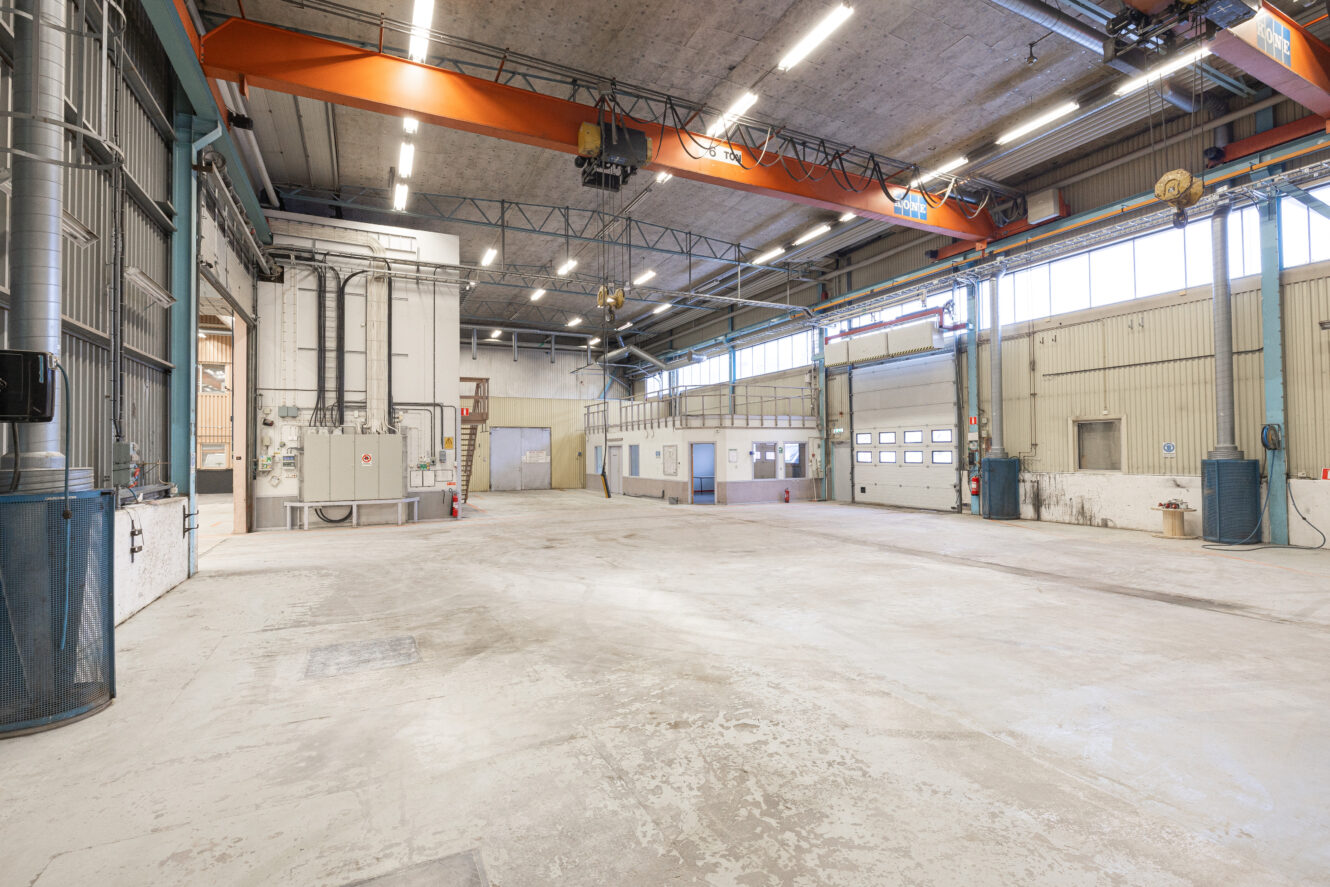 Stor, tom industrilokal med högt i tak, traverskranar, stora fönster och flera garageportar. Golv och väggar visar tecken på slitage.