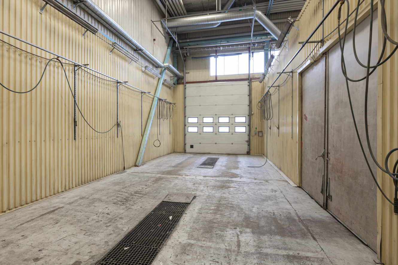 Tom industritvätthall med väggar av korrugerad metall, rörledningar, hängande slangar, golvbrunnar och en stängd garageport i bortre änden.