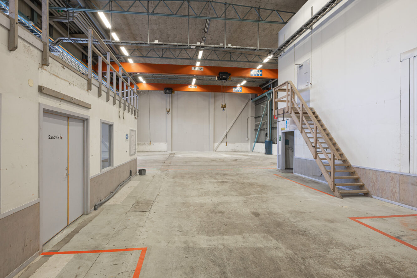 Rymlig tom industrihall med betonggolv, högt i tak, trätrappa och markerade ytor på marken.