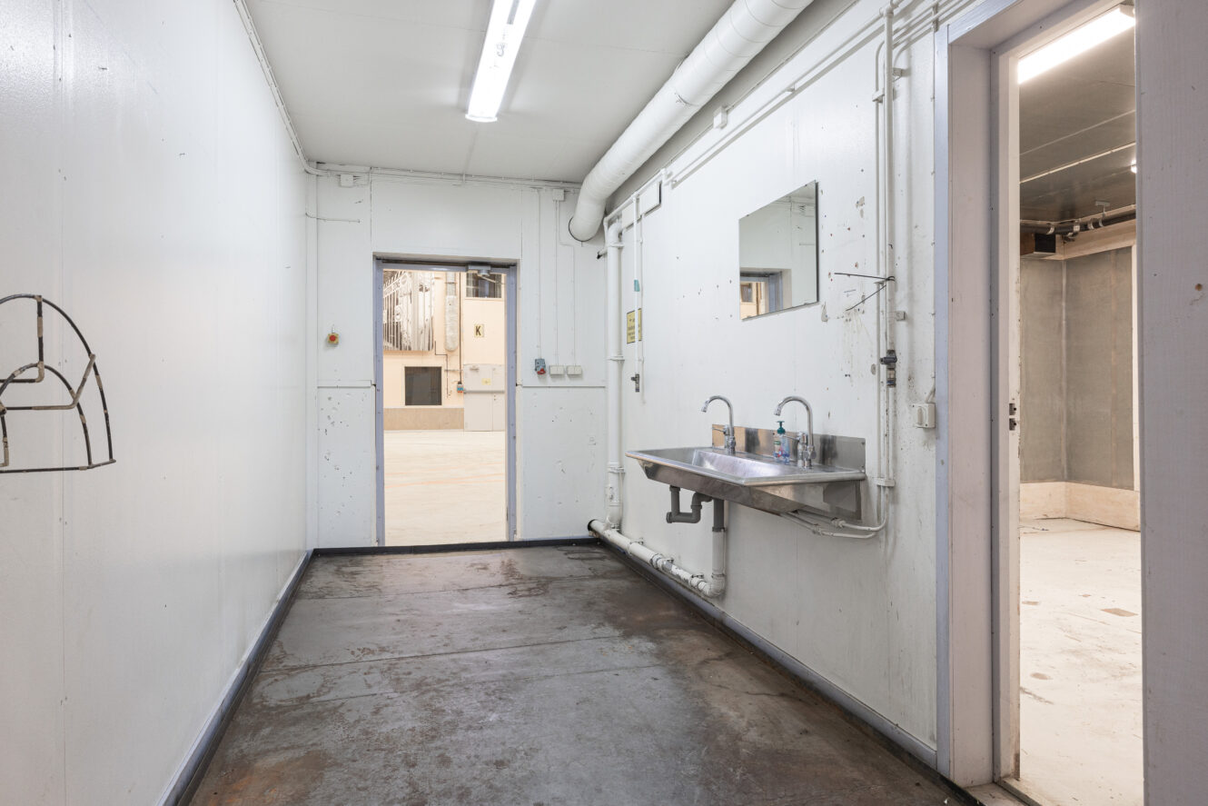 Industriellt tvättrum med diskbänkar och speglar i metall, vita väggar, synliga rör och lysrörsbelysning, anslutet till intilliggande rum med betonggolv.