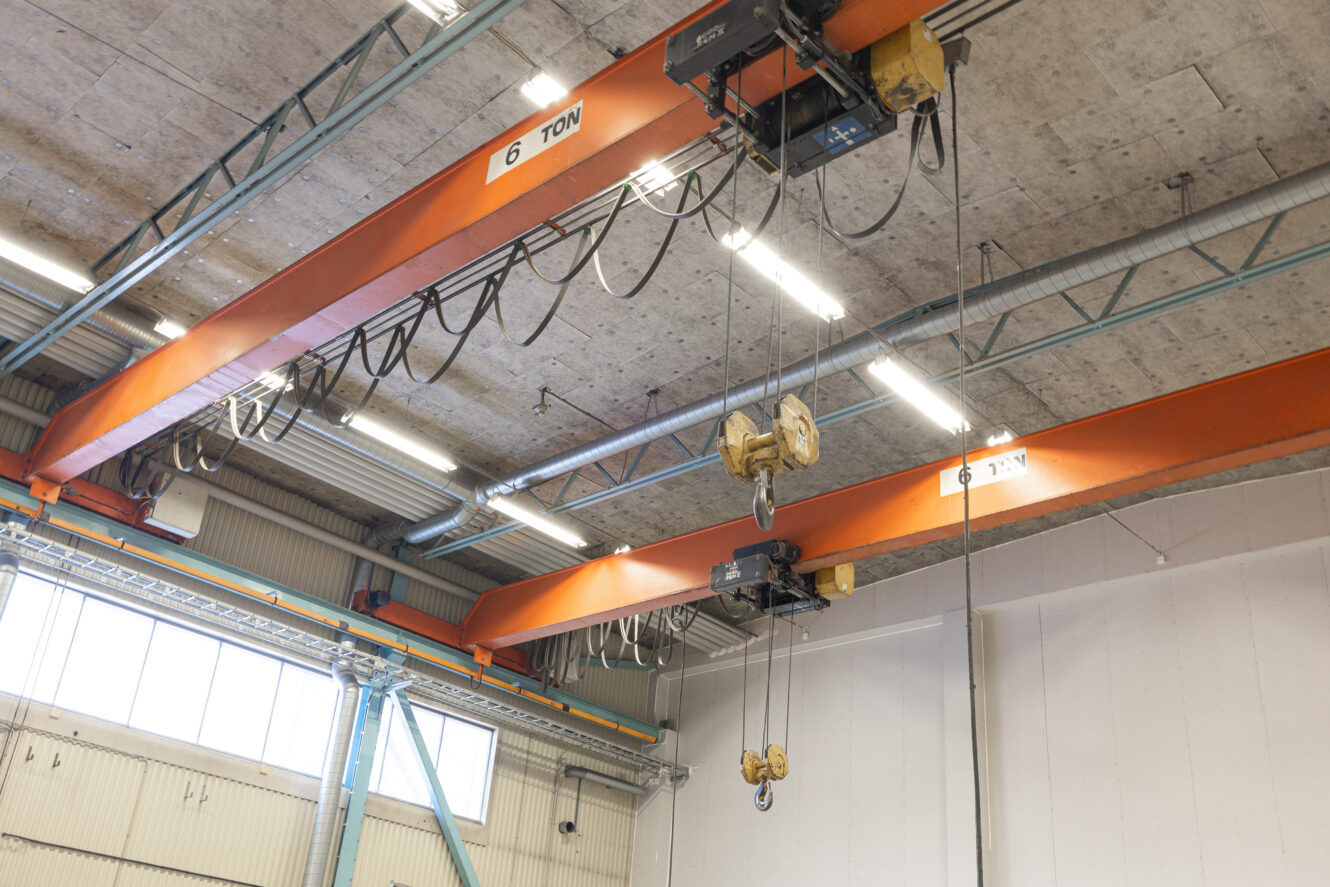 Orange traverskranar med 6 tons kapacitet hänger från taket i en industrilagerlokal, med kablar och krokar hängande nedåt.