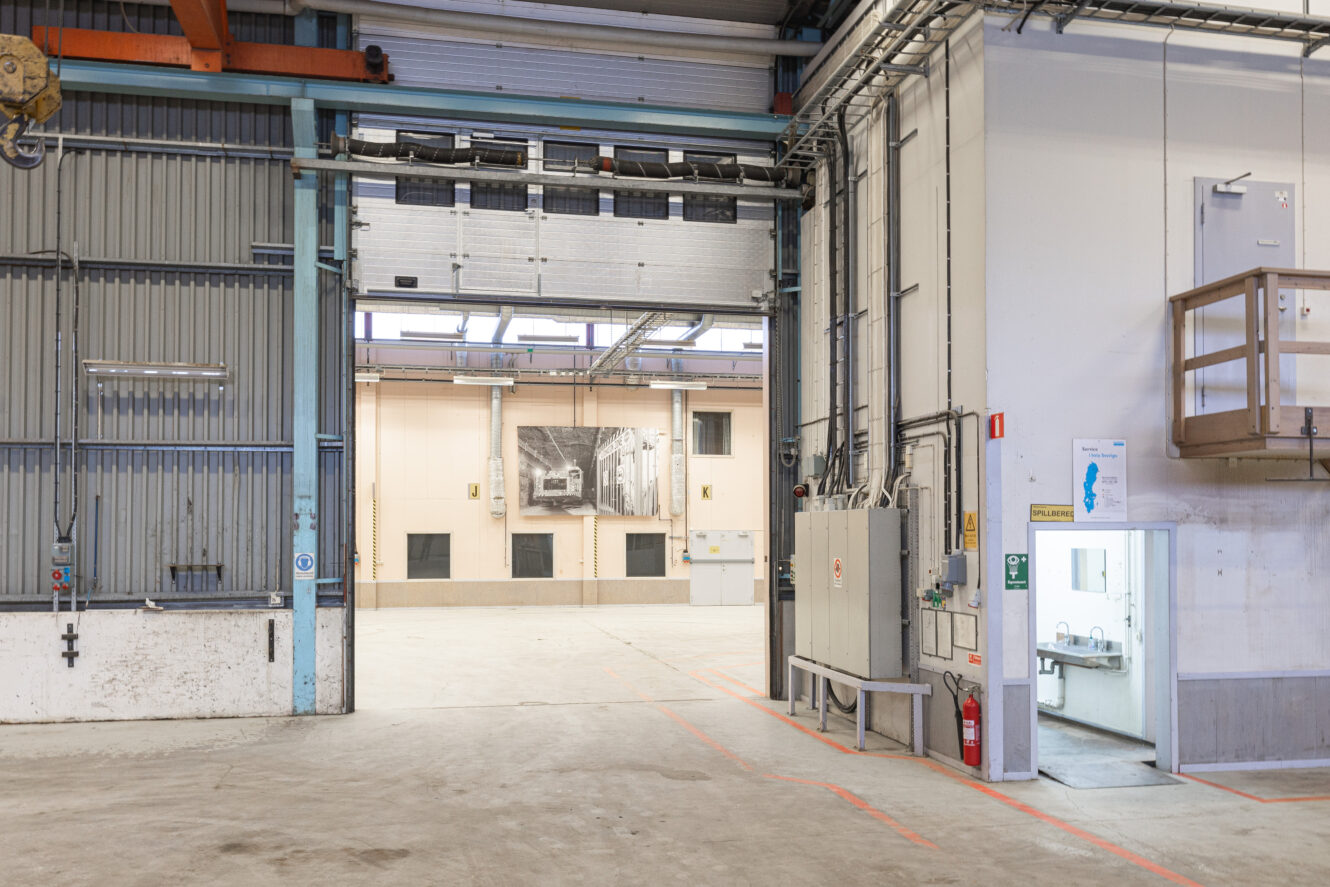 Interiör av ett tomt industrilager med en delvis öppen garageport, synliga kontrollpaneler, brandsläckare och ett litet rum till höger.