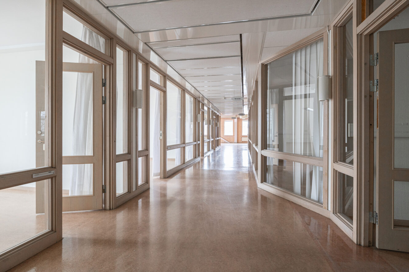 En ren, tom korridor i en byggnad med öppna trädörrar och stora glasfönster längs båda sidor, upplyst av naturligt ljus.