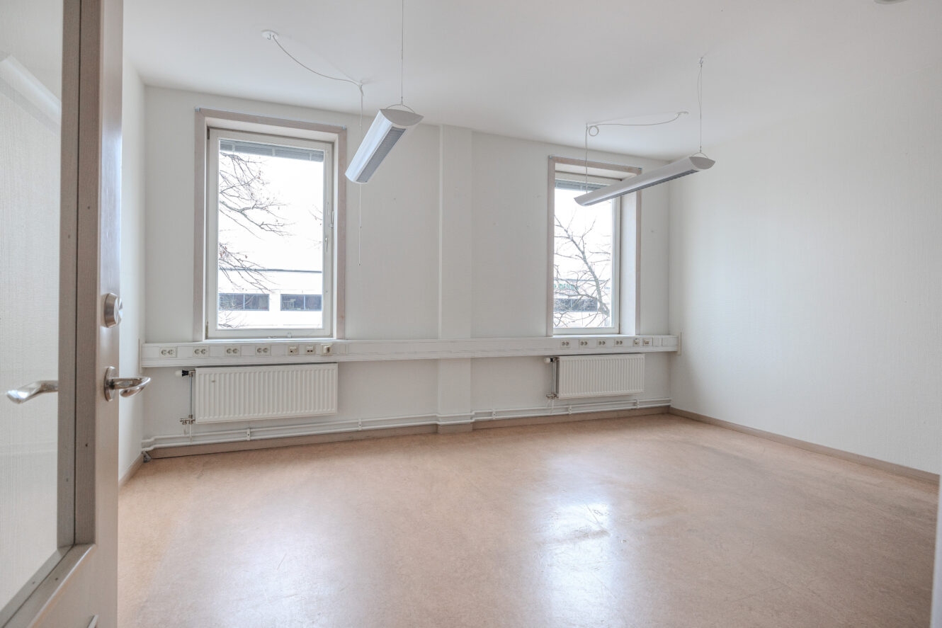 Tomt, omöblerat rum med två stora fönster, vita väggar, beige golv, taklampor och synliga radiatorer under fönstren.