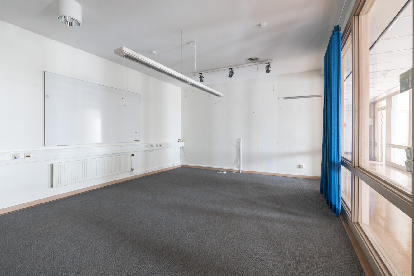 Tomt rum med grå matta, vita väggar, en stor whiteboard, taklampor, eluttag, blå gardiner och stora fönster på höger sida.