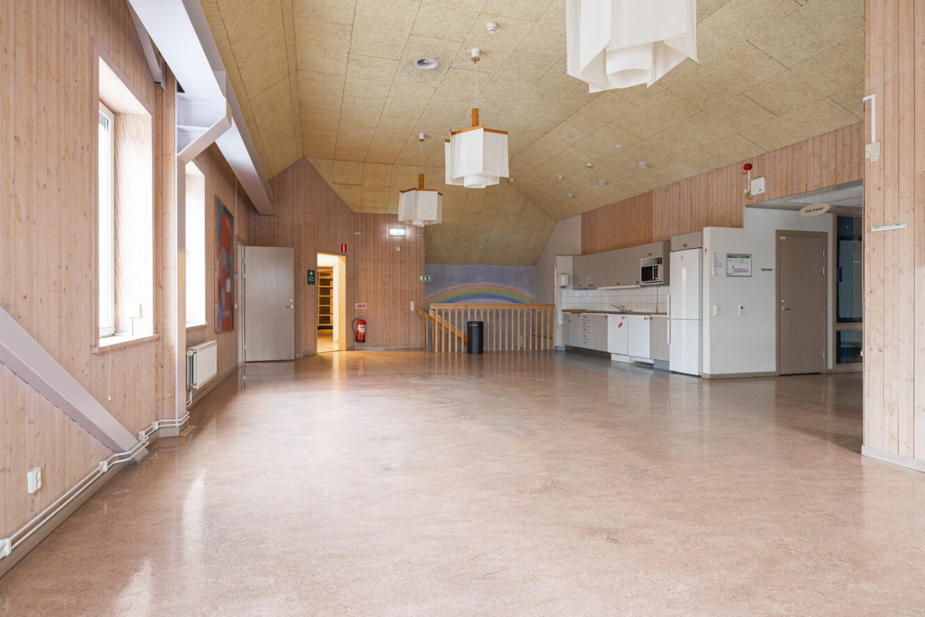 Ett rymligt, tomt rum med träpanel på väggarna, ett pentry till höger, hängande armaturer och en regnbågsmuralmålning i bakgrunden.