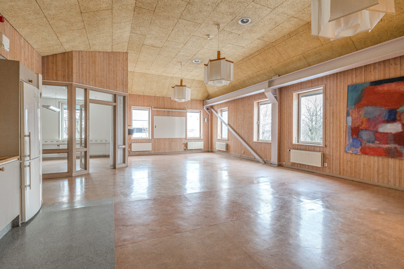 Ett rymligt, tomt rum med väggar och tak i trä, stora fönster, whiteboardtavlor, ljusarmaturer, ett pentry och en färgstark abstrakt målning på ena väggen.