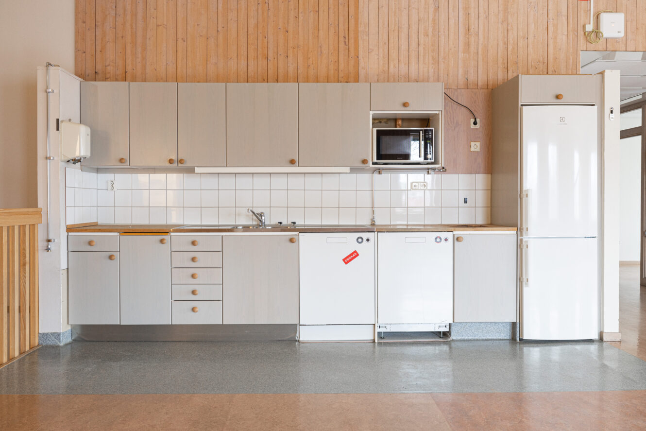 Ett minimalistiskt kök med skåp i ljust trä, vit kakelplatta, inbyggda vitvaror, en mikrovågsugn och ett kylskåp mot en vägg med träpanel.