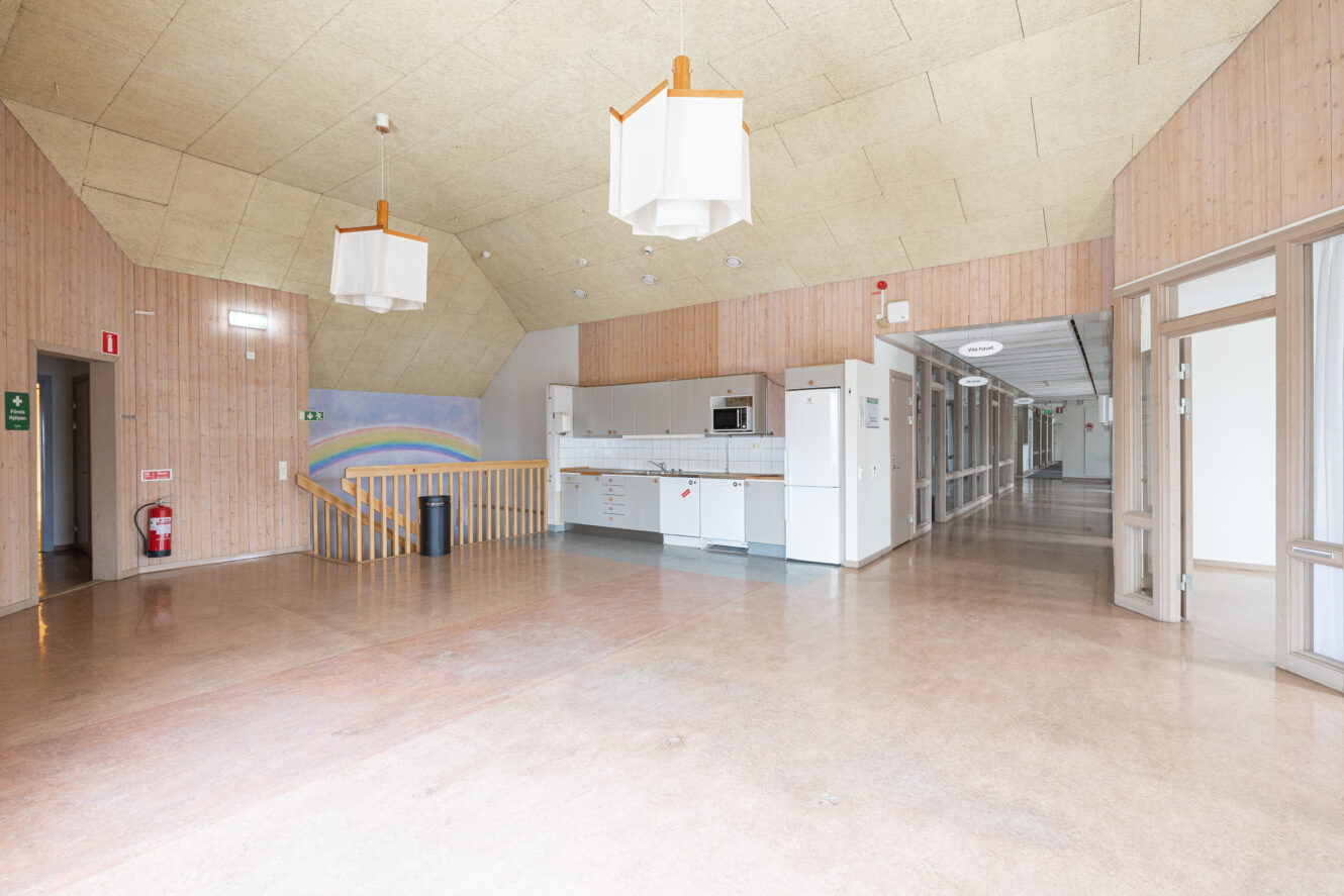 Rymlig, tom korridor med träpanel på väggarna, pentry, taklampor och stora fönster; en regnbågsmuralmålning på ena väggen.