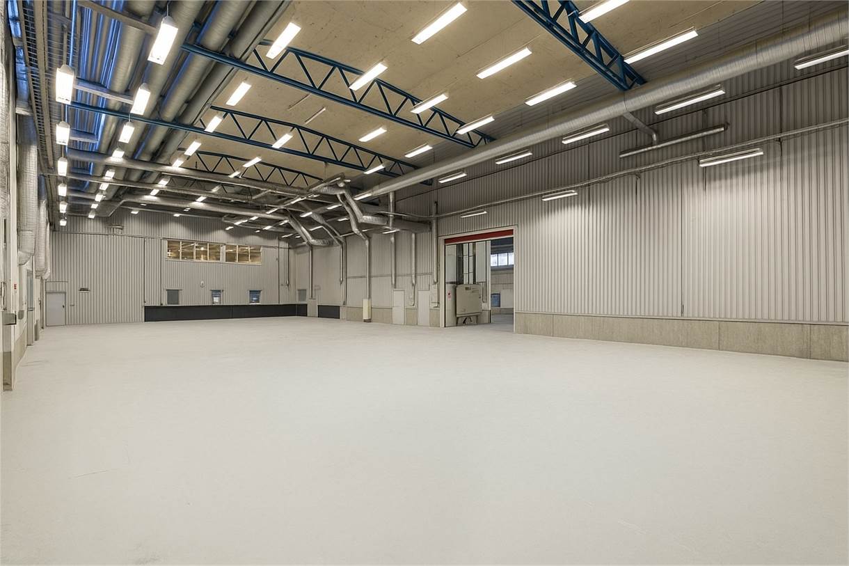 Rymlig industrilagerlokal med högt i tak, synliga kanaler, stora fönster och stark takbelysning. Golvet är tomt och utrymmet ser rent och modernt ut.