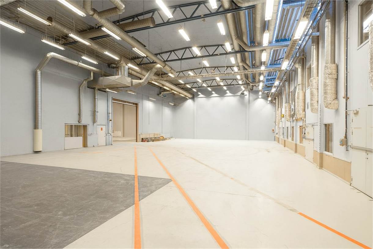 Stort, tomt industrilager med högt i tak, takbelysning, synliga kanaler och orangefärgade golvlinjer; väggarna är ljusgrå med några lådor och pallar nära baksidan.