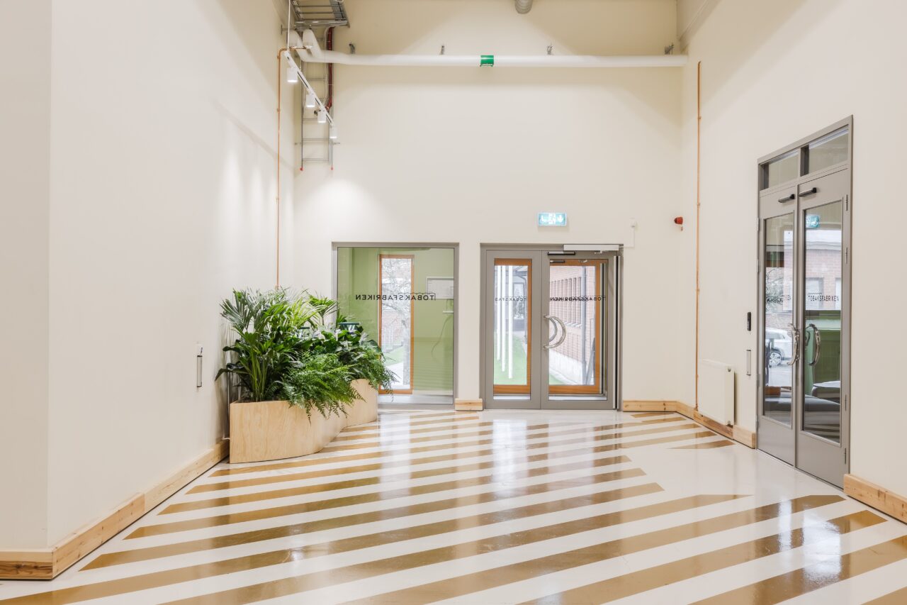 En ljus, modern hall med randigt golv, krukväxter till vänster, dubbeldörrar i glas i slutet och en vägg med fönster till höger.