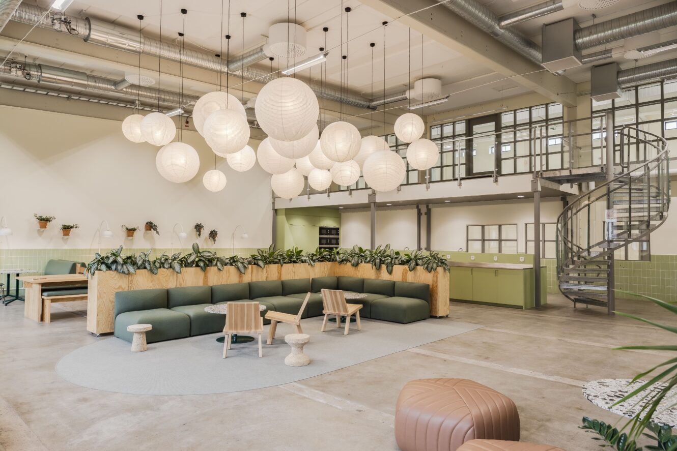 Modern kontorslounge med gröna soffor, trästolar, hängande globlampor, spiraltrappa och inomhusväxter i ett rymligt, öppet område med högt i tak och stora fönster.