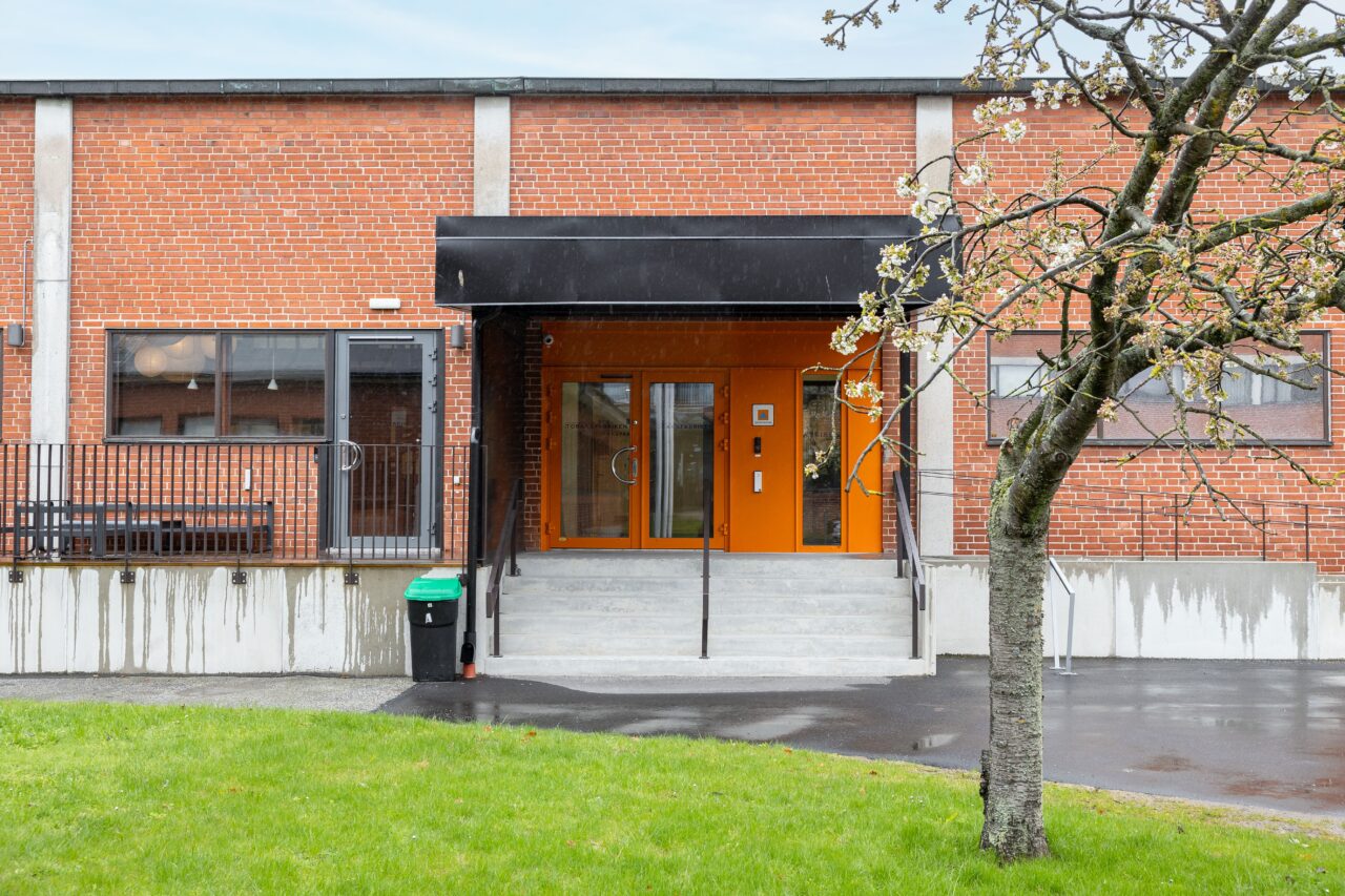 En tegelbyggnad med orange entré och betongtrappa, svart markis, metallräcken och ett litet träd framför.