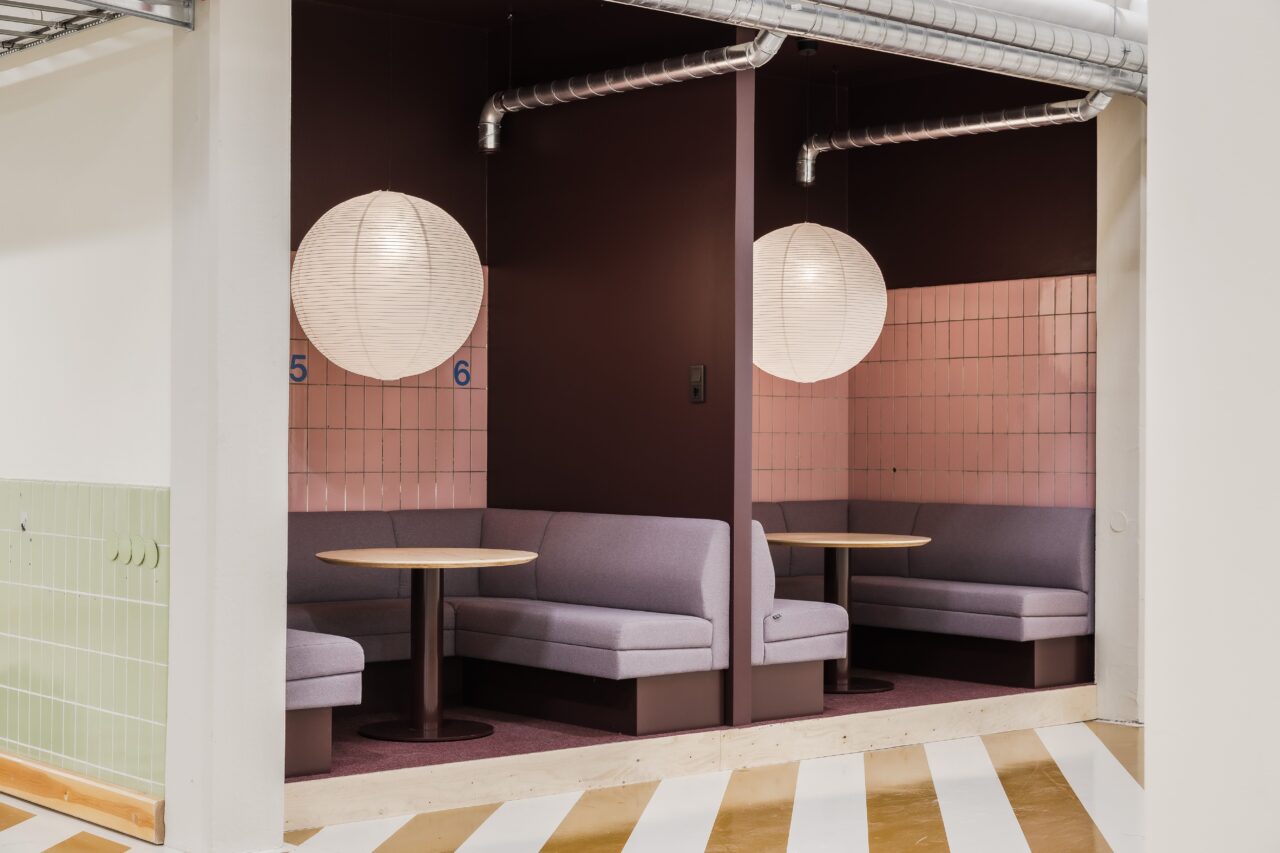 Två halvprivata bås med runda bord och sittkuddar skiljs åt av en vägg, var och en upplyst av en vit papperslykta, i en modern inomhusmiljö.