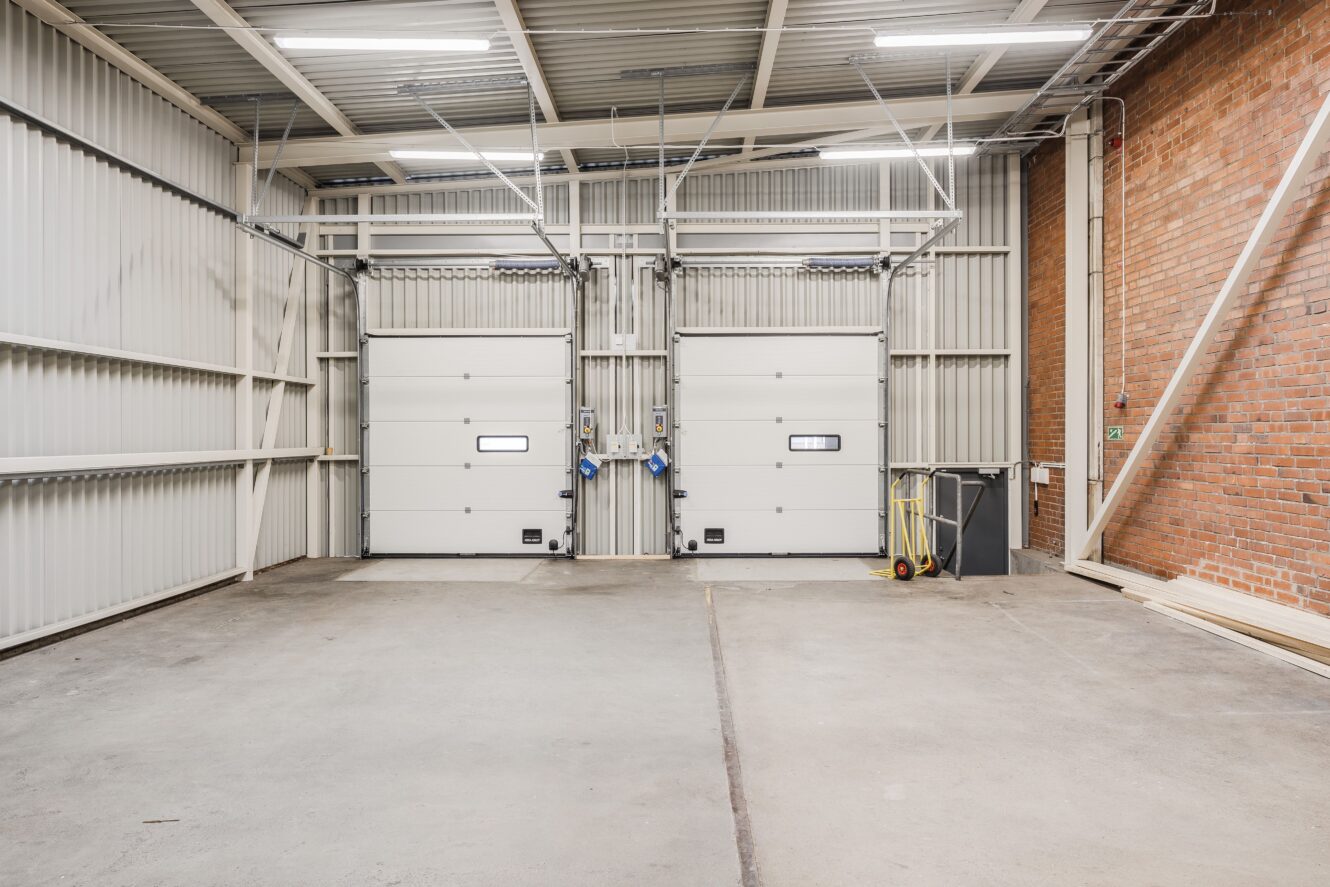 Industriell lagerlokal med två stängda vita garageportar, synlig tegelvägg till höger och rengöringsutrustning i hörnet.