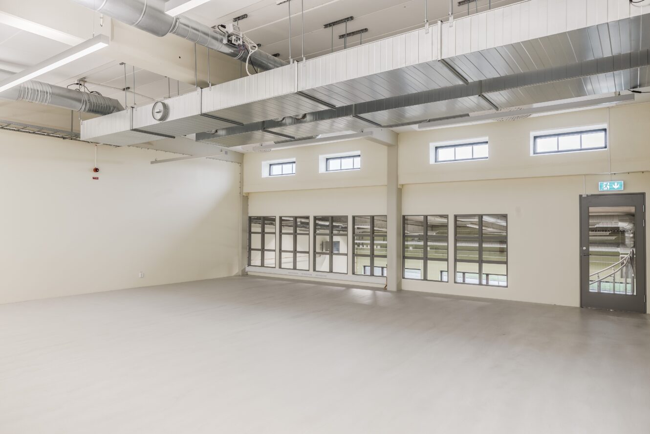 Tomt rum i industriell stil med synliga ventilationsrör, högt i tak, stora fönster och en grå dörr som leder till ett trapphus.
