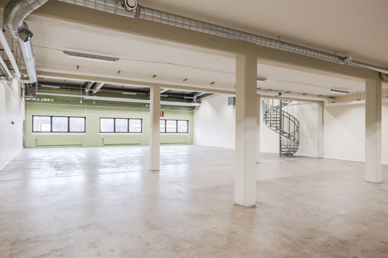 Rymlig, tom industrilokal med stora fönster, synliga kanaler, betonggolv och en spiraltrappa i metall i det bakre hörnet.